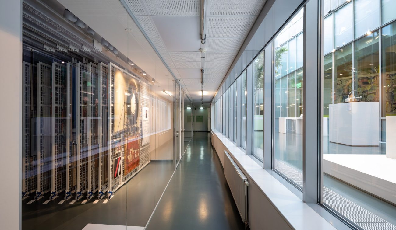 Glazen corridor in SCHUNCK Heerlen met rechts uitzicht op het atrium en links achter glas uitschuifbare rekken met ingelijste kunstwerken; strak, modern interieur met veel daglicht.