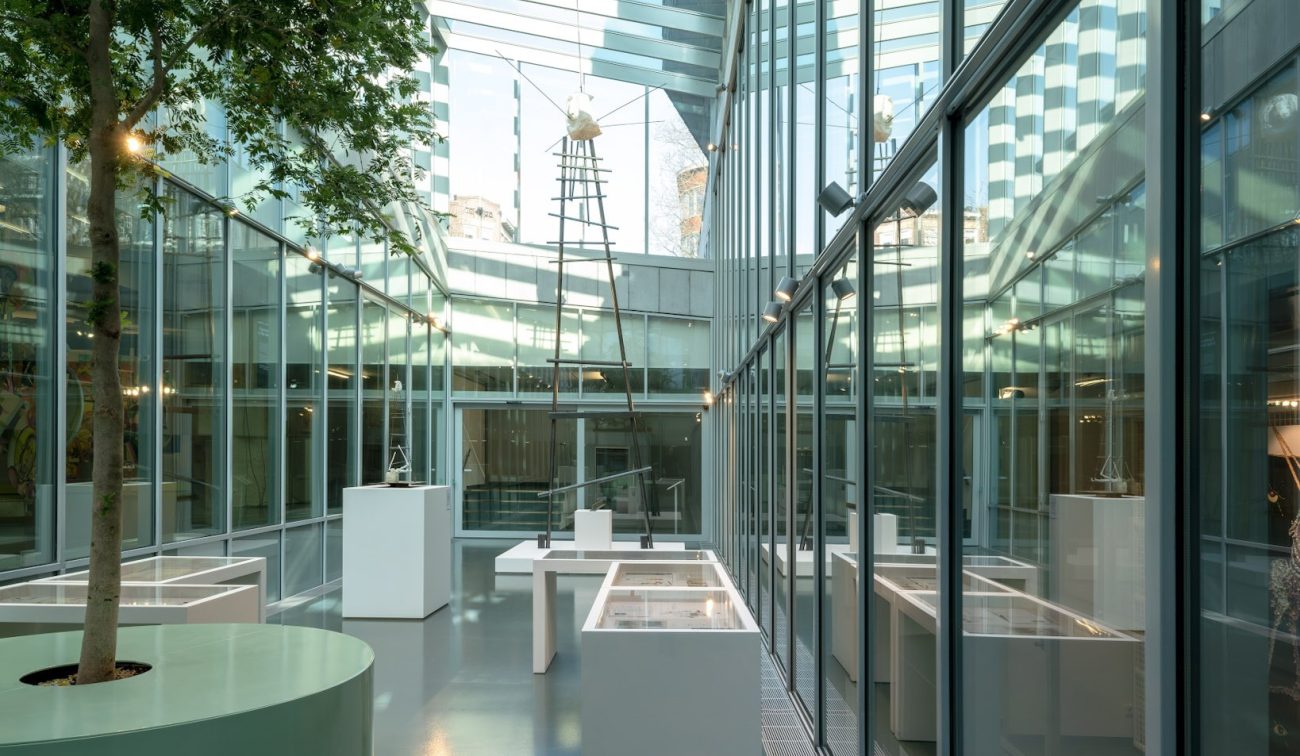Licht glazen atrium van SCHUNCK Heerlen met glazen dak, slanke staal-/glasgevels, witte vitrinekasten en sokkels, en een ronde plantenbak; reflecties benadrukken de transparante, moderne transformatie.