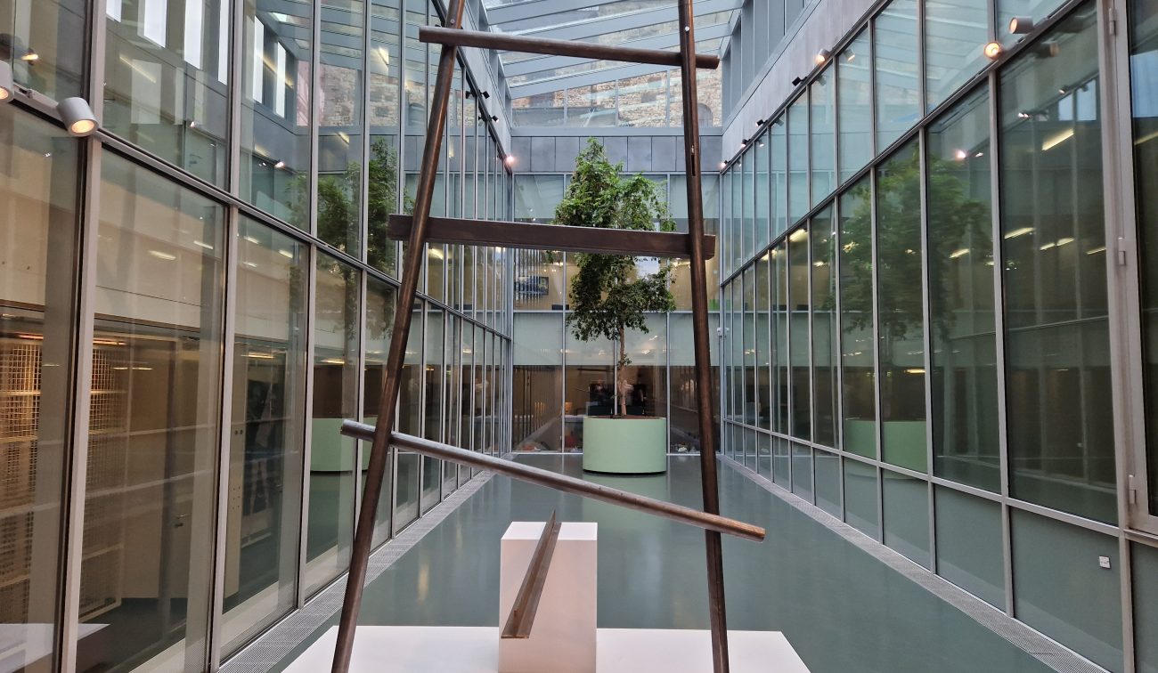 SCHUNCK Heerlen – glazen atrium met sheddak, centraal een stalen ‘ladder’-sculptuur op sokkel en aan het eind een boom in ronde groene plantenbak, geflankeerd door glazen gevels.