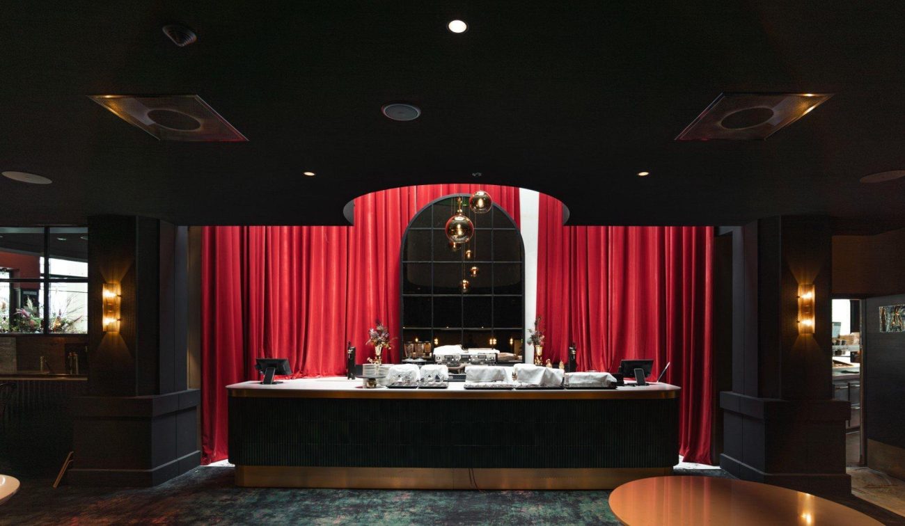 Foyerbalie met rode fluwelen gordijnen, boogvormige spiegel en hanglampen in een donker interieur.