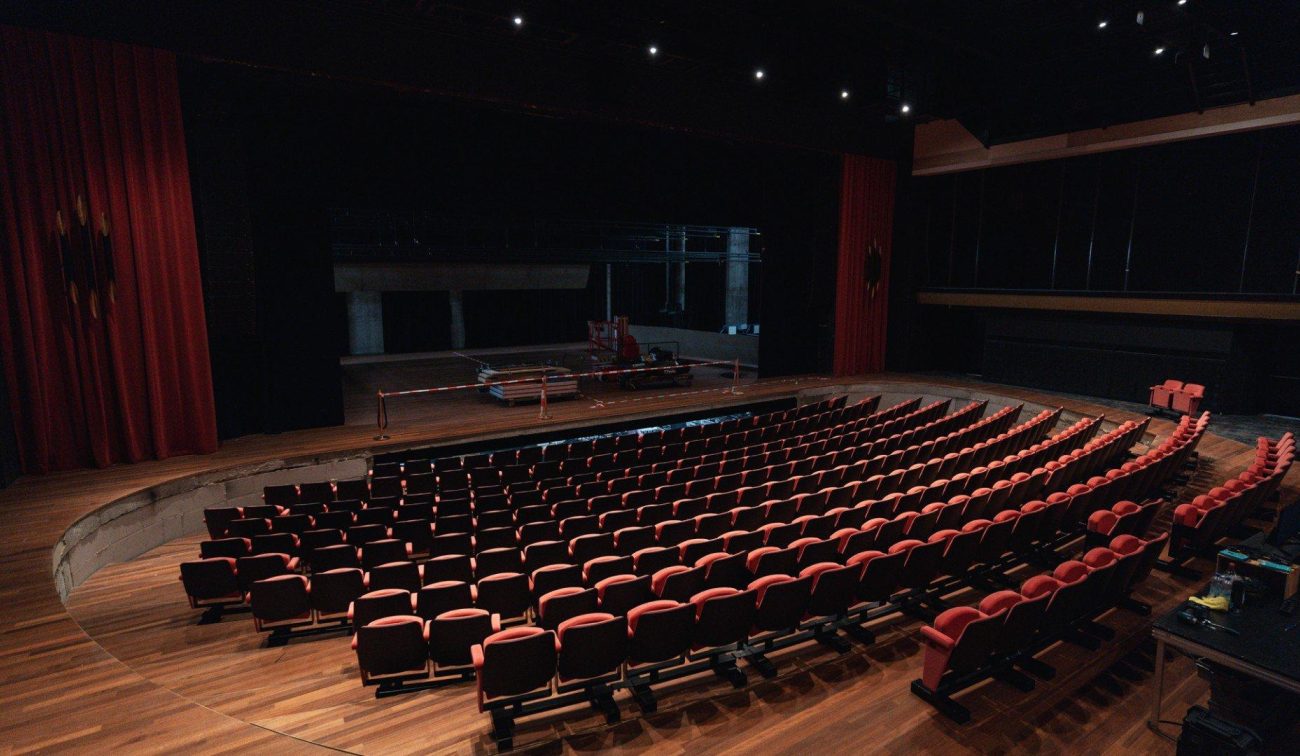 Theaterzaal in verbouwing met gebogen rijen rode stoelen, houten vloer en podium met bouwmaterialen; rode gordijnen en gedimde verlichting in Theaterhotel De Oranjerie, Roermond.