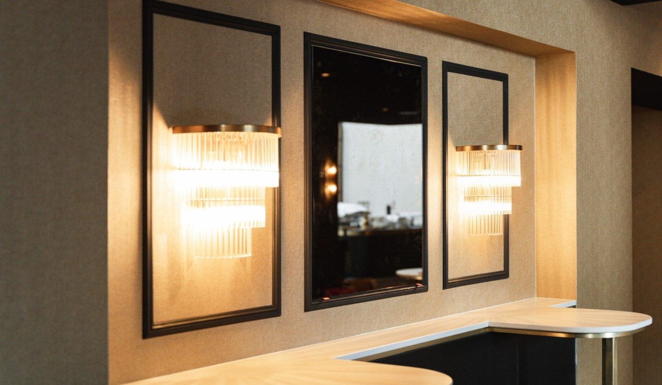 Detail foyer Theaterhotel De Oranjerie – twee glazen wandlampen met warme gloed naast een zwarte spiegel in panelenwand boven een afgeronde bar.