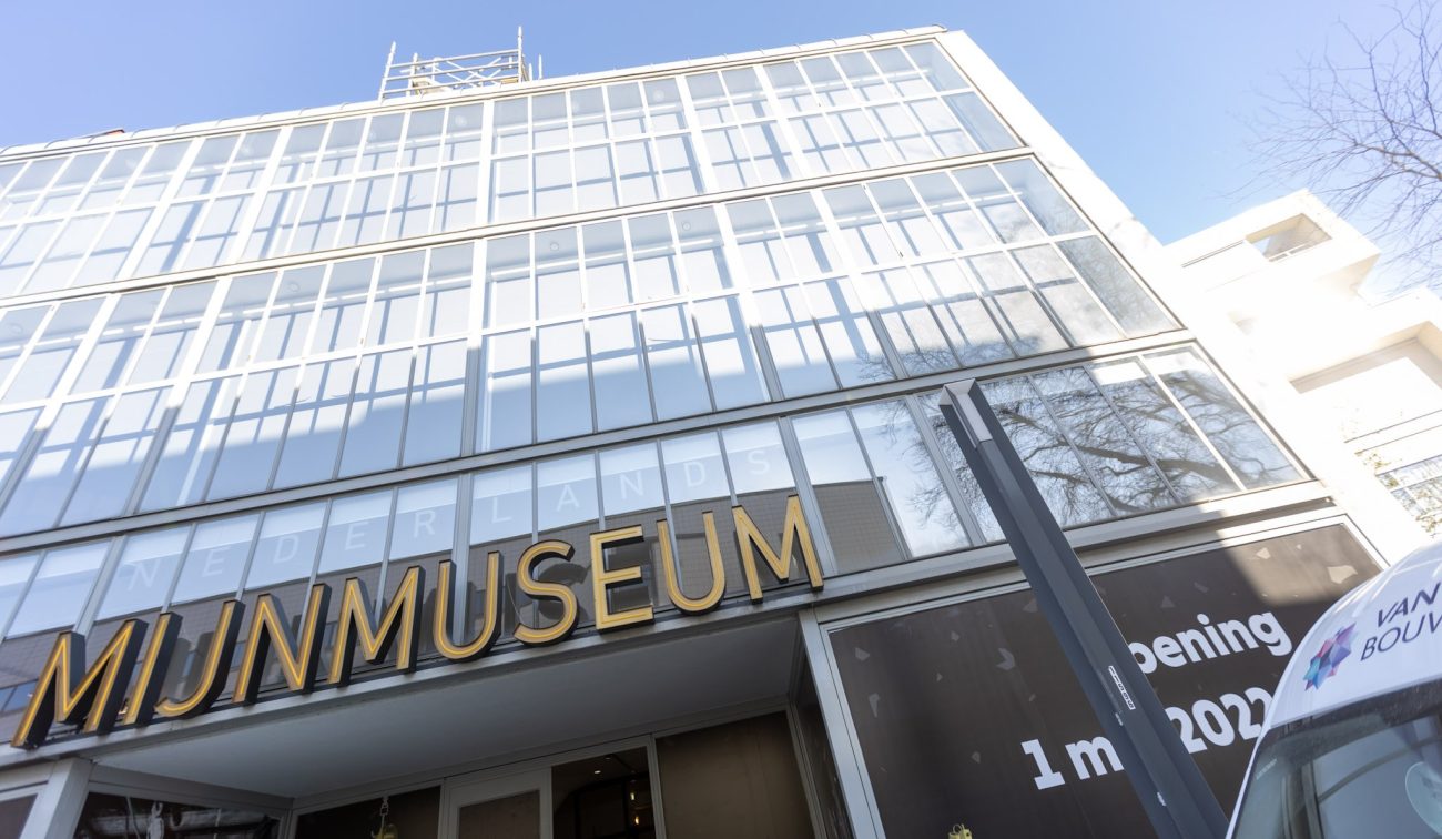Glazen gevel van het Nederlands Mijnmuseum in Heerlen met grote gouden letters ‘MIJNMUSEUM’, steigerwerk bovenaan en raamstickers met ‘Opening 1 mei 2022’; rechts de neus van een bedrijfsbus.