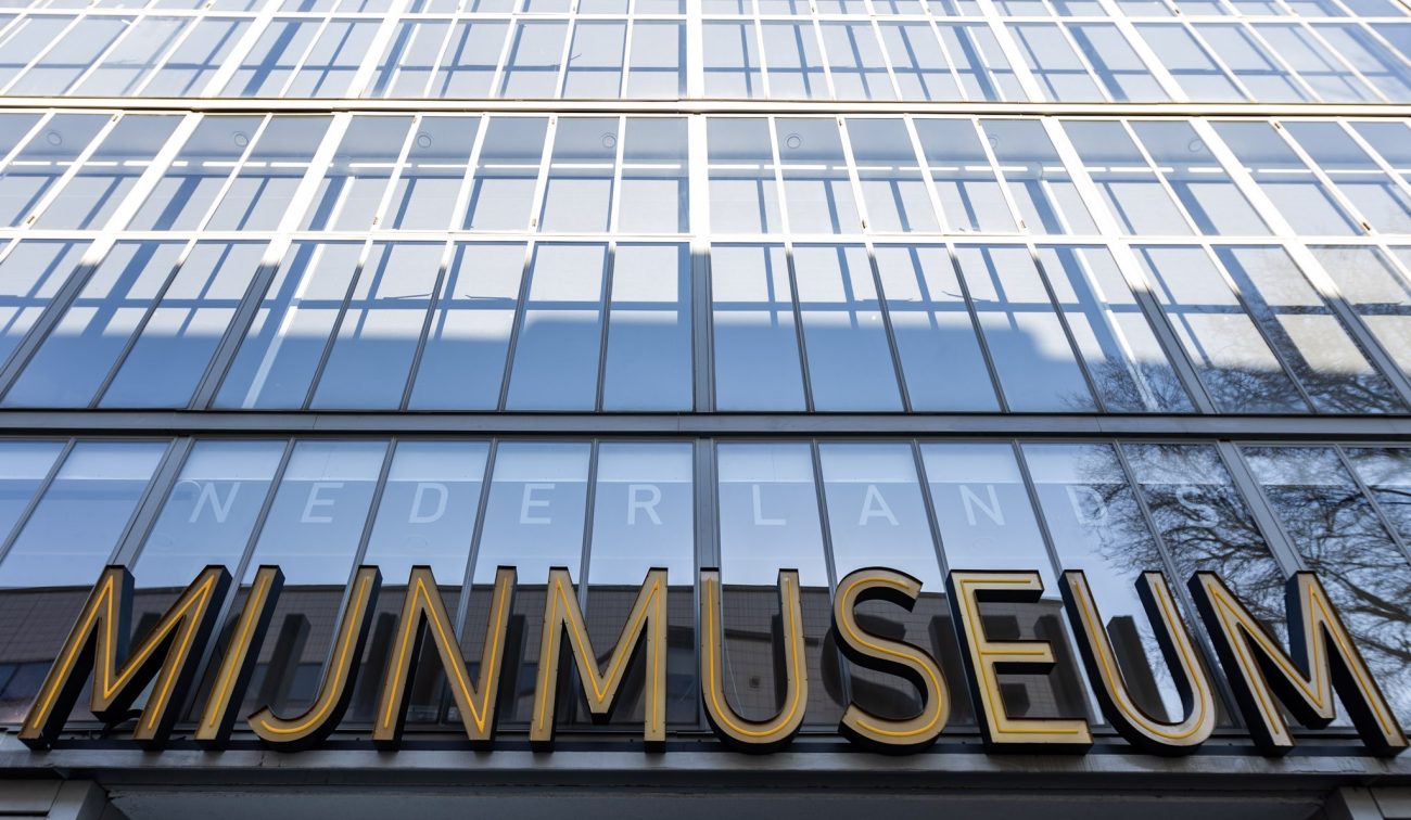 Close-up van de glazen rastergevel van het Nederlands Mijnmuseum in Heerlen met grote gouden letters ‘MIJNMUSEUM’ en weerspiegelingen van lucht en bomen.