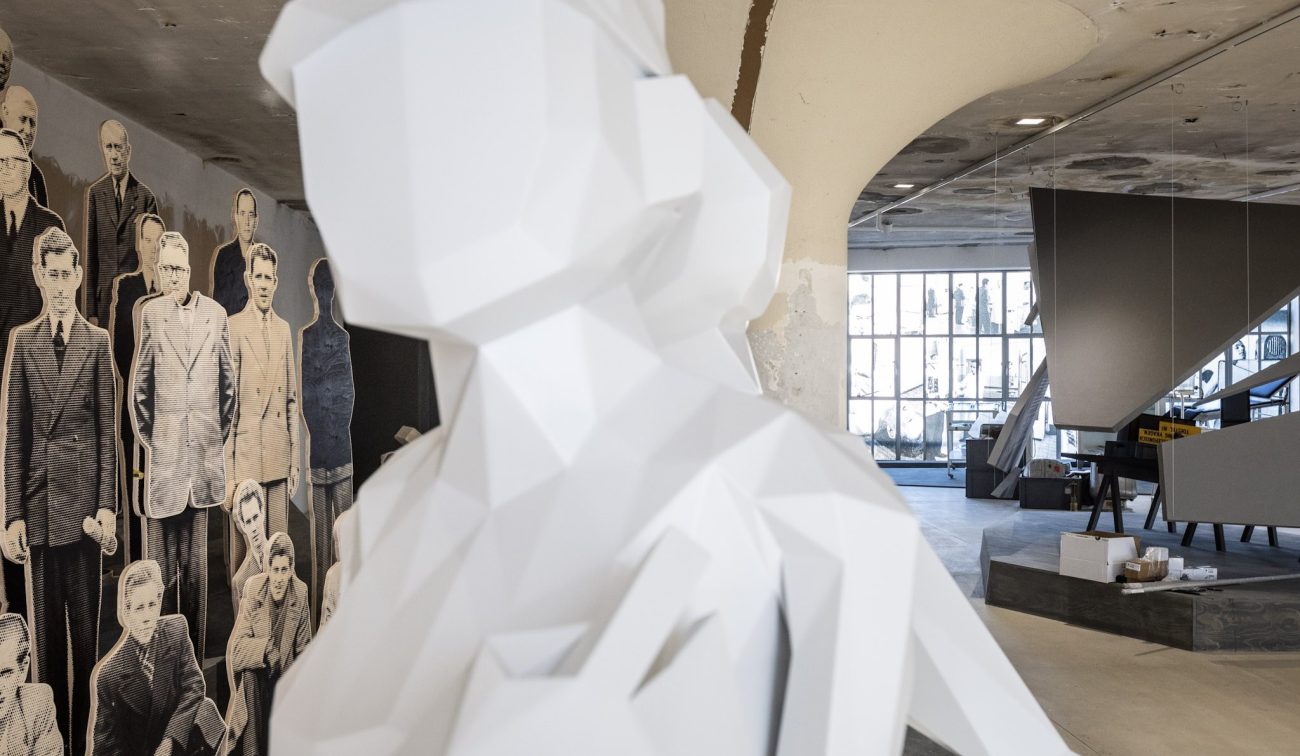 Voorgrond een witte, facetvormige sculptuur; op de achtergrond de in verbouwing zijnde zaal van het Nederlands Mijnmuseum met paddenstoelkolommen, grote industriële ramen en levensgrote portretten van mijnwerkers tegen de muur.