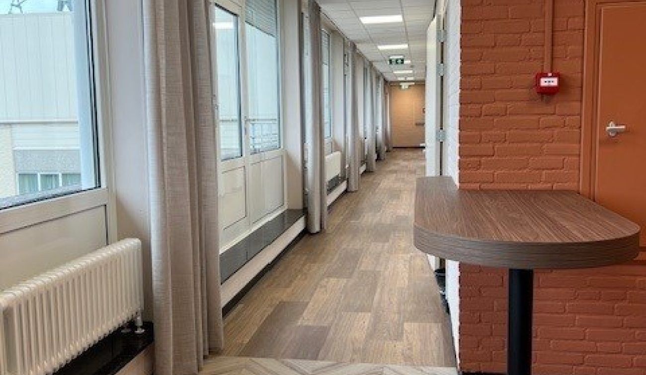 Renovatie Schuttershof Brunssum – lichte gang met visgraat- en plankvloer, bakstenen wand, hoge ramen en gordijnen