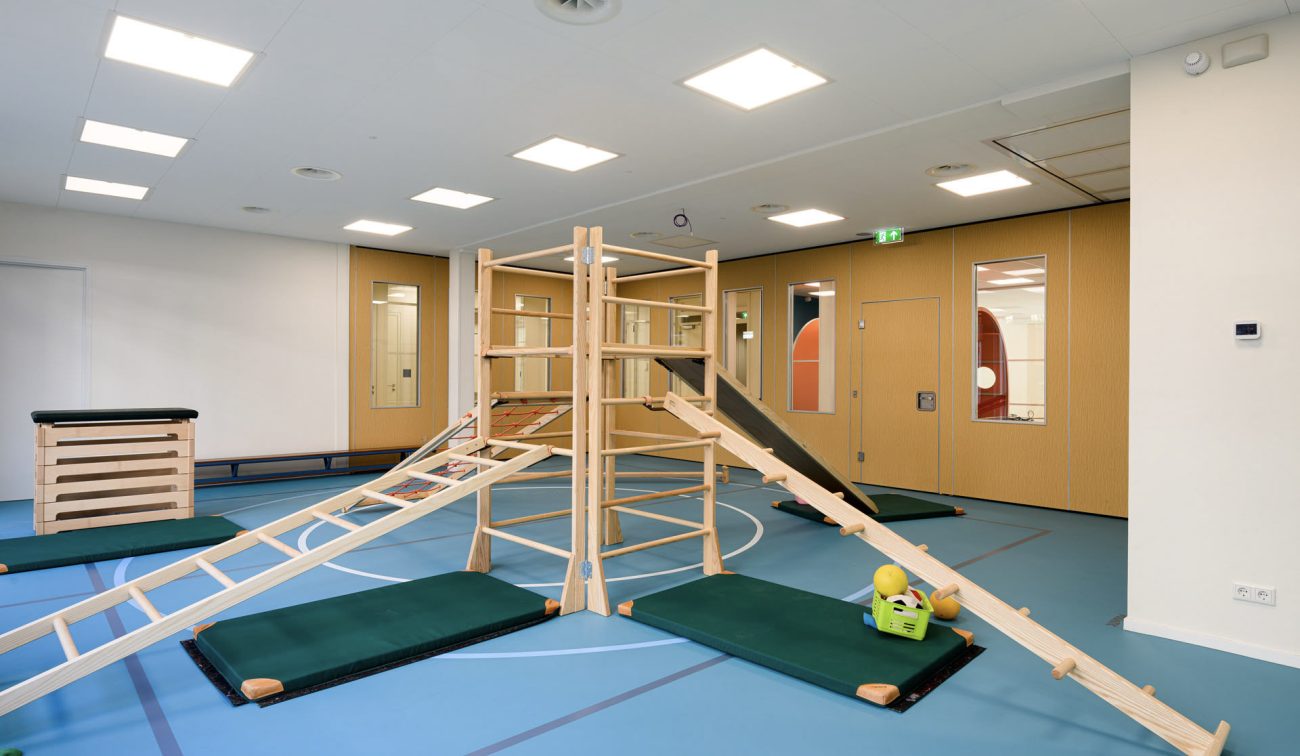Renovatie Kindcentrum LEF Utrecht – speels/veilig gymlokaal met dempende sportvloer, houten klimtoestellen en zichtvensters naar aangrenzende ruimtes.