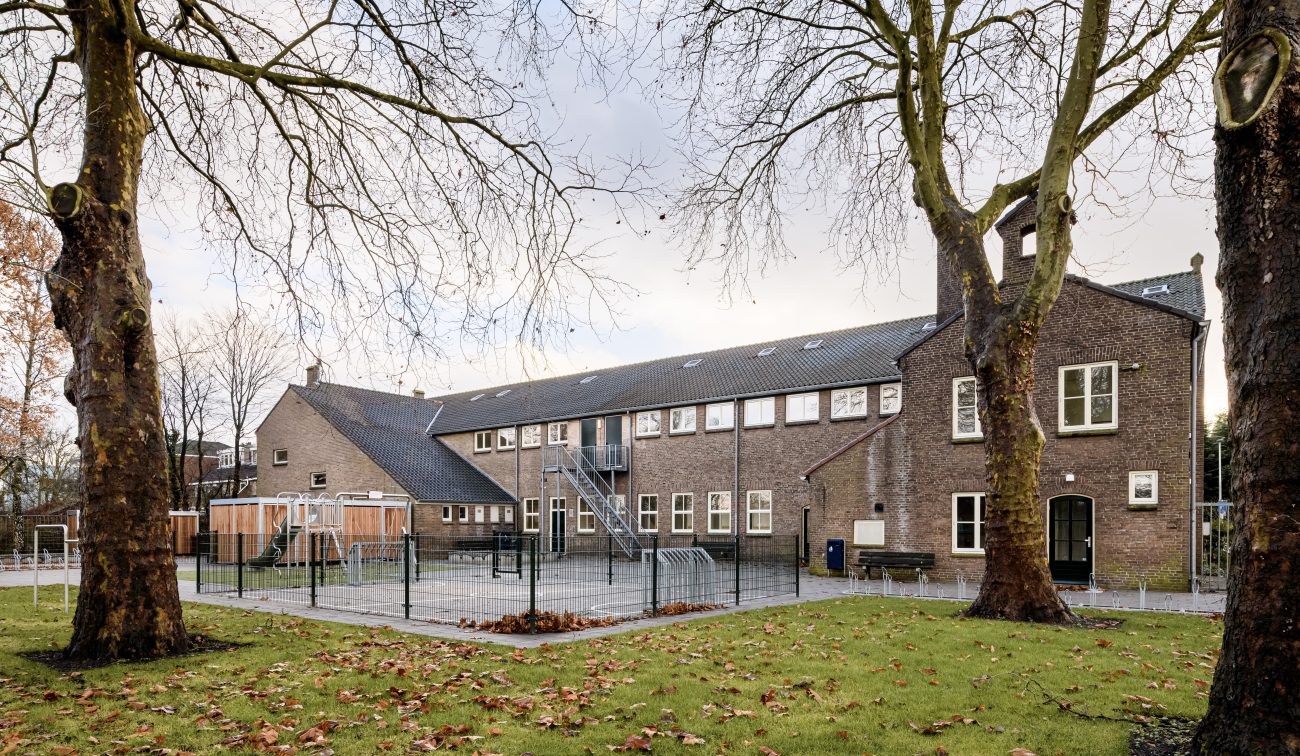 Gerestaureerd schoolgebouw in baksteen met speelplaats, hekwerk en twee grote platanen op voorgrond.