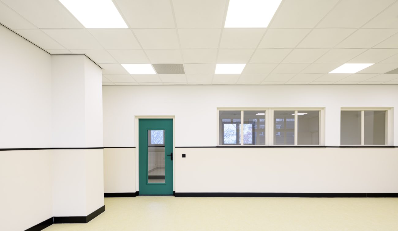 Strakke schoolruimte; halfhoge lambrisering, turkoois binnendeur, raamstrook naar gang en lichte vloer.