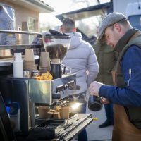 Man bereidt koffie bij een mobiele koffiekar terwijl mensen eromheen staan.