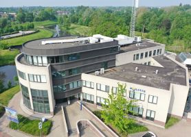 Luchtfoto van het politiebureau in Roermond met ronde glazen hoek en donkere bovenbouw, zicht op platte daken met installaties en het ‘POLITIE’-logo aan de gevel, omringd door groen en water.