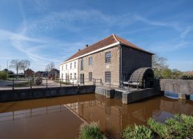Grathemermolen in Grathem – bakstenen watermolen met rood pannendak en groot waterrad aan het water, met spiegeling in de molenvijver onder blauwe lucht.