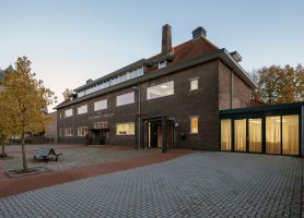 Gerestaureerd schoolgebouw St. Gerardus Majella in Heerlen met donkerbruine bakstenen gevel en rood pannendak, vernieuwde entree en moderne glazen uitbouw met gordijnen, gezien vanaf het betegelde schoolplein met boom en bankje in avondlicht.