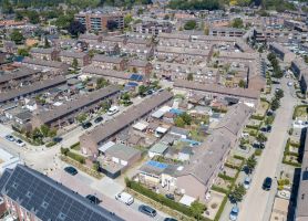 Luchtfoto van een dichtbebouwde woonwijk met lange rijen rijtjeshuizen, smalle straten met geparkeerde auto’s, achtertuinen en enkele daken met zonnepanelen; links is een bouw- of renovatieplaats zichtbaar.