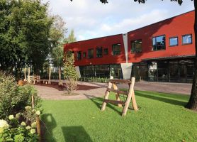 Groen schoolplein met gazon en natuurlijke speeltoestellen van hout; op de achtergrond de rood beklede gevel van basisschool Den Doelhof met grote ramen en glazen entree.