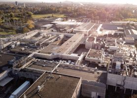 Luchtfoto van de uitgebreid en gerenoveerd ogende Mora-fabriek in Maastricht, met een uitgestrekt daklandschap vol installaties, leidingen en stoompluimen, omringd door groen en bedrijfsgebouwen.