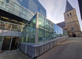 Exterieur SCHUNCK Heerlen – glazen corridor/uitbouw met stalen kozijnen naast de historische kerk met toren, gezien vanaf het pleintje.