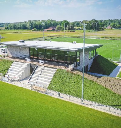 Luchtfoto van het moderne clubgebouw van DVO’20 in Kelpen-Oler, met glazen pui, tribunetrap en groene taluds, omgeven door meerdere voetbalvelden.