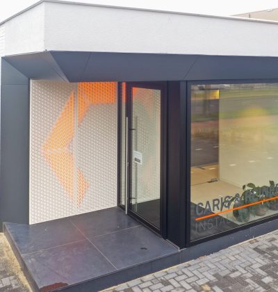 Hoekentree van gerenoveerd kantoor Caris & Reiner in Sittard met zwart kader, glazen pui en geperforeerde wand met oranje verlichting op een donkere tegelopstap.