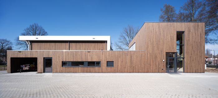 : Voorgevel van de nieuwbouw Brandweerpost Sittard-Oost met lange, horizontale houtbeklede volumes, een open garagebox links, smalle raamstrook en hoge glazen entree rechts onder een strakblauwe lucht.