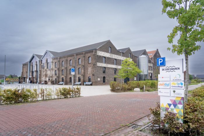 Gerestaureerde Greswarenfabriek in Reuver met bakstenen gevel, grote glazen entree en historische opdruk ‘GEBR. TEEUWEN’; voorplein met bankjes, fietsrekken en een jonge boom.