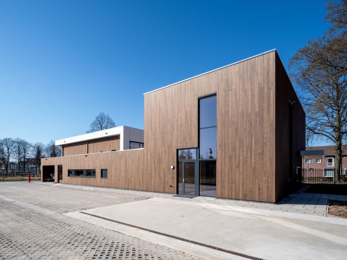 Dagopname van de nieuwe brandweerpost Sittard-Oost met houtbeklede gevel, hoge glazen entree en wit volume erachter, aan een ruim verhard voorterrein onder een strakblauwe lucht.