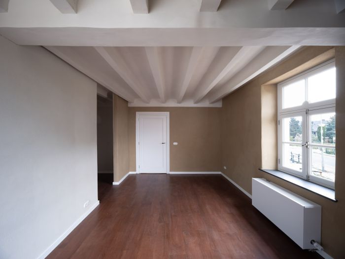 Gerestaureerde kamer in de Grathemermolen met wit balkenplafond, leemkleurige wanden, houten vloer, witte paneeldeur en diep venster met radiator en daglicht.
