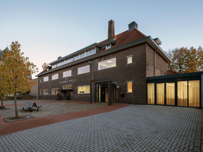 Gerestaureerd schoolgebouw St. Gerardus Majella in Heerlen met donkerbruine bakstenen gevel en rood pannendak, vernieuwde entree en moderne glazen uitbouw met gordijnen, gezien vanaf het betegelde schoolplein met boom en bankje in avondlicht.
