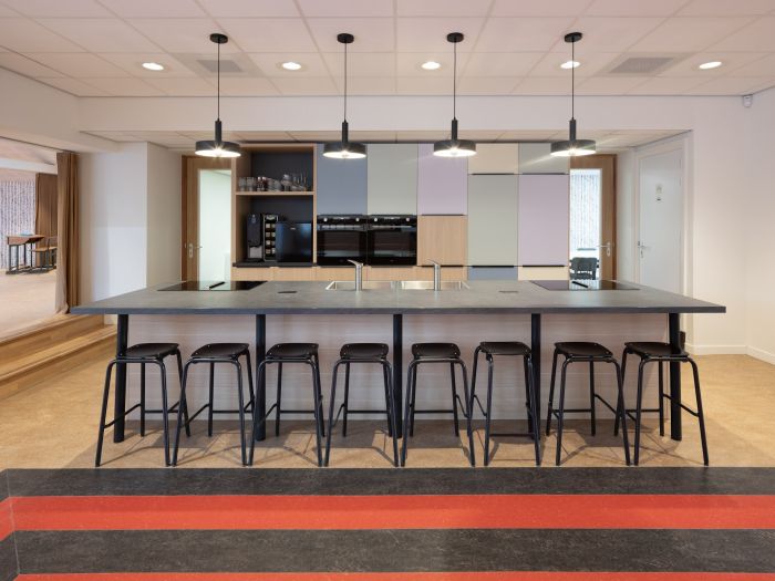 Kook/eetruimte Majella-school met groot kookeiland, acht zwarte barkrukken, vier hanglampen en pastelkleurige kastenwand met inbouwovens en koffiecorner.