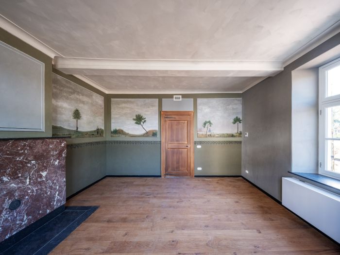 Gerestaureerde kamer in de Grathemermolen met houten vloer, groen-grijze wanden met landschappelijke muurschilderingen, houten deur centraal, marmeren schouw links en groot raam rechts.