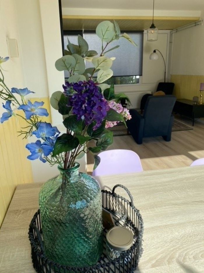 Woonkamer Staatsliedenbuurt Tilburg – close-up van groene glazen vaas met kleurrijke bloemen op tafel, met zitgedeelte en staande lamp op de achtergrond.