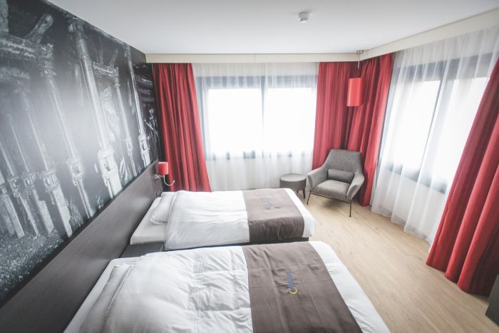 Interieur hotelkamer Geleen – twee bedden, rode overgordijnen, sheer vitrage en grijze stoel met bijzettafel