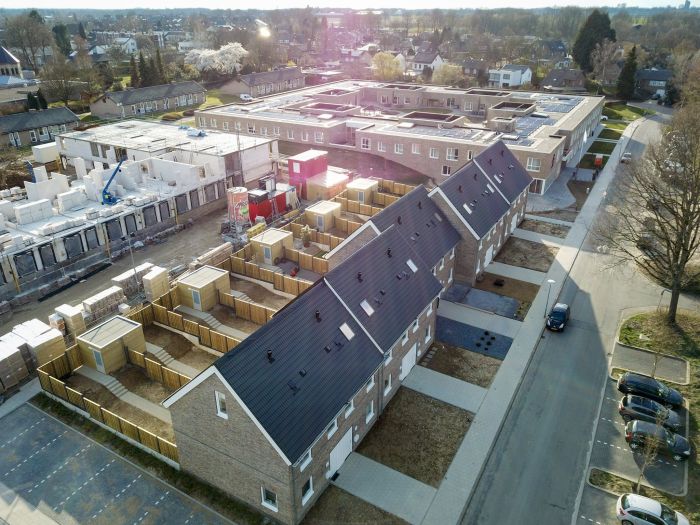Luchtfoto nieuwbouw sociale eengezinswoningen in Hulsberg met rijwoningen met zwarte zadeldaken, achtertuinen en naastgelegen bouwplaats met appartementen in aanbouw.