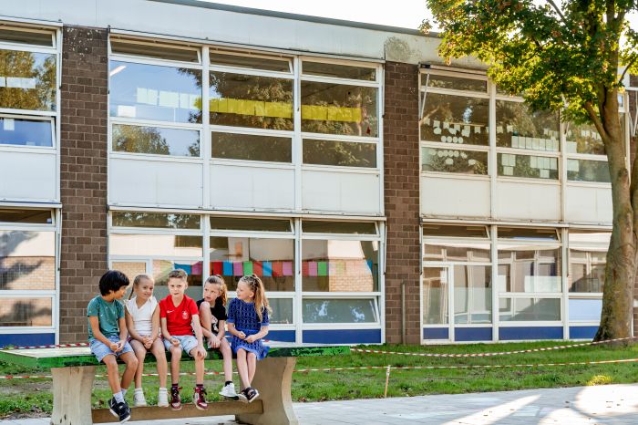 Kinderen op schoolplein – groepje op bankje voor bakstenen schoolgebouw met grote ramen en kleurige slingers