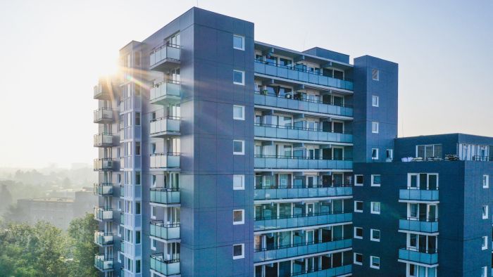 Close-up van de gerenoveerde Aambosveldflat in Heerlen met grijze gevel en glazen balkons; ochtendzon met lensflare schijnt langs de hoek van de woontoren.