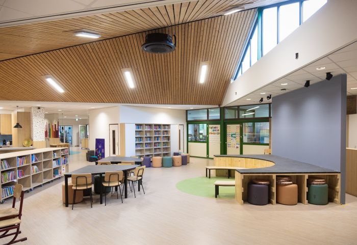 Open leerplein met houten plafond, boekenkasten en werktafels; rechts een halfrond podium met zitpoefs en groene accenten bij grote ramen.