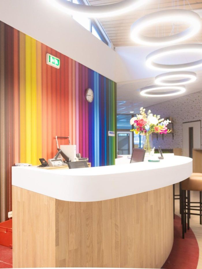 Receptiebalie met witte, afgeronde top en houten front; op de achtergrond een regenboogkleurige wand met klok en ronde ringlampen, vaas met bloemen op de balie.