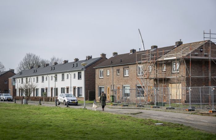 ij woningen in Maastricht; links gerenoveerde witte gevels, rechts bakstenen huizen in steigers. Een persoon met hond loopt langs en er staat een auto geparkeerd.