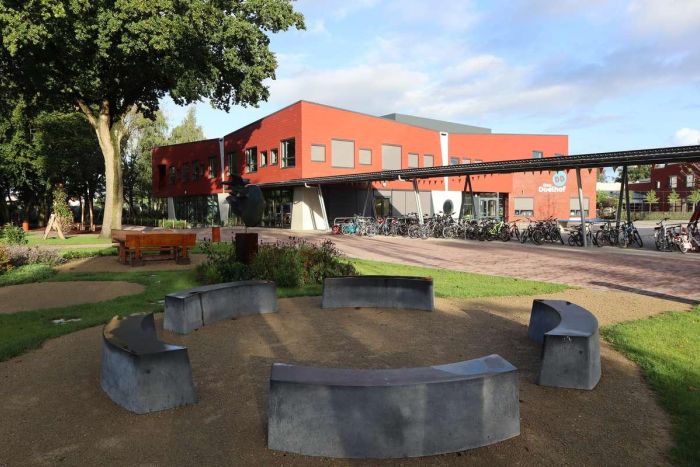 Nieuwbouw basisschool Den Doelhof in Meijel met rood gevelvolume en luifel; fietsenstalling met veel fietsen langs de gevel. Op de voorgrond een ronde zitplek van betonnen banken in de groene schooltuin met bomen.
