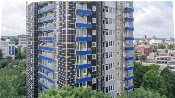 Close-up van een hoge woontoren in renovatie met blauwe balkonbanden en zwarte rastergevel, steigers langs de gevel en de stad op de achtergrond.