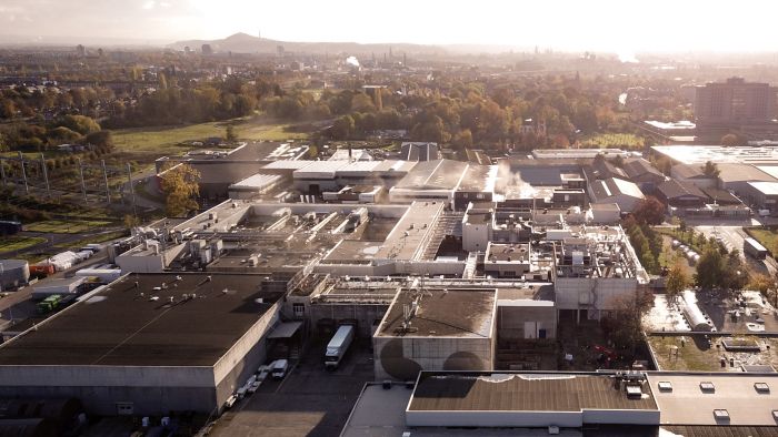 Luchtfoto van de uitgebreide en gerenoveerde Mora-fabriek in Maastricht, met daken vol installaties en stoompluimen, laadtrucks op het terrein en herfstkleurige bomen rondom.