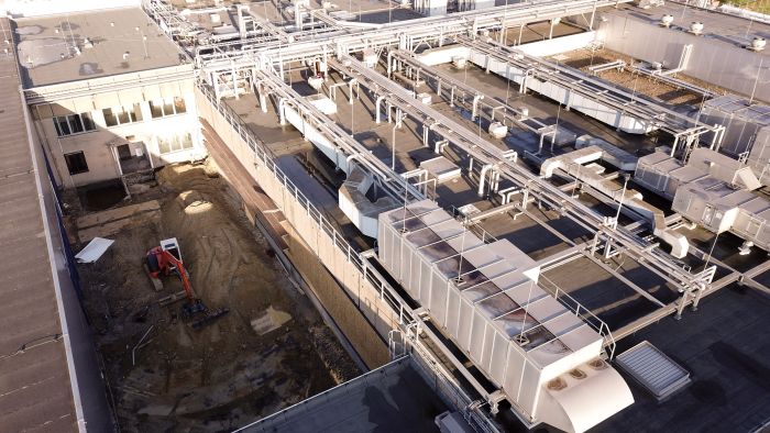 Schuine luchtfoto Mora-fabriek Maastricht met een smalle bouwkuip naast het gebouw; linksonder een rode graafmachine in het zand, rechts grote daken vol leidingen en HVAC-kanalen in de zon.