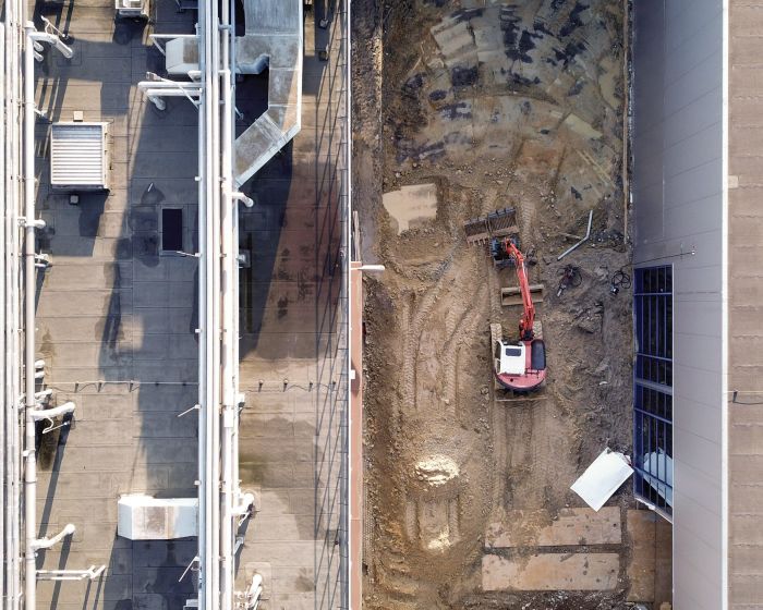 Luchtfoto bouwsleuf bij Mora-fabriek Maastricht: smalle doorgang tussen twee gebouwen met een rode graafmachine in het midden, duidelijk zicht op rups- en graafsporen; links dak met leidingen, rechts metalen gevel met raamstroken.