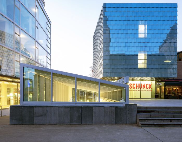 SCHUNCK Heerlen – buitenaanzicht met schuine glazen corridor/lichtstraat tussen het glazen hoofdgebouw en de met zink beklede toren; bij de entree het rode ‘SCHUNCK’ logo zichtbaar.