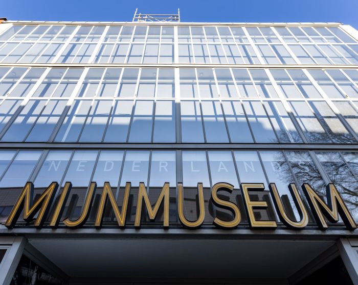 Close-up van de glazen rastergevel van het Nederlands Mijnmuseum in Heerlen met grote gouden letters ‘MIJNMUSEUM’ en weerspiegelingen van lucht en bomen.