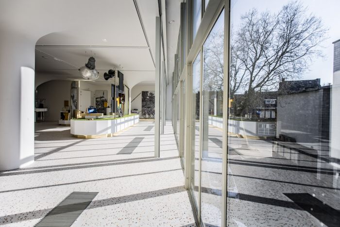Lichte museumruimte met grote glazen gevel en lange schaduwen op een terrazzo vloer; in het midden een lang wit vitrinedisplay met mijngereedschap. Buiten zijn bomen en gebouwen zichtbaar.