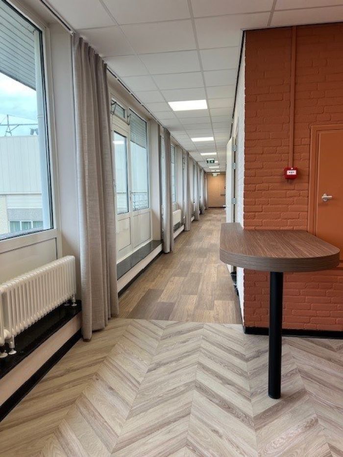 Renovatie Schuttershof Brunssum – lichte gang met visgraat- en plankvloer, bakstenen wand, hoge ramen en gordijnen