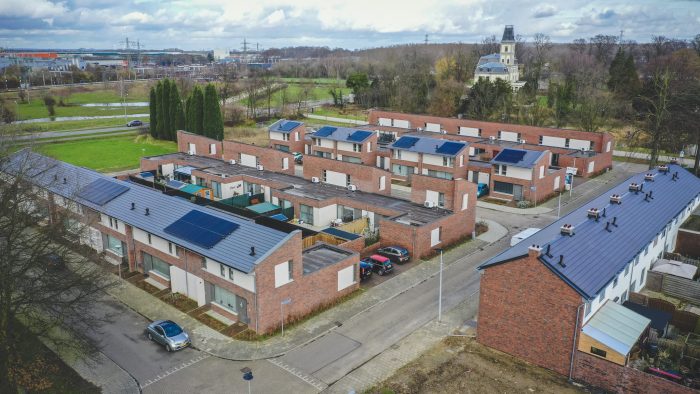 Schuine luchtfoto van nieuwbouwwijk Nazareth (Maastricht): rode bakstenen rijwoningen met zonnepanelen, binnentuinen en geparkeerde auto’s; groenstroken en een kerktoren op de achtergrond.