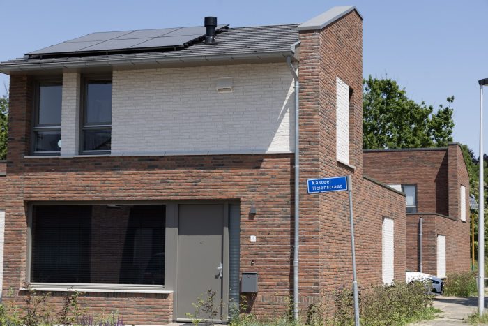 Hoek van een moderne bakstenen woning met witte accenten en zonnepanelen op het grijze dak; grijze voordeur en raam aan de straatzijde, met een blauw straatnaambord ‘Kasteel Heilenstraat’ op de hoek.
